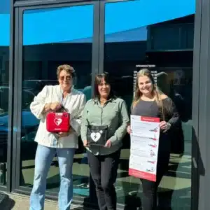 Drei Personen stehen vor Gebäude mit AED und Aufsteller